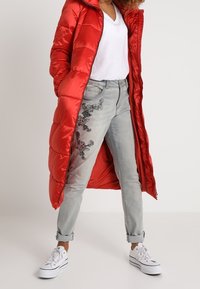 Rød pufferjakke, lang, shiny materiale, båret over en hvid V-hals skjorte. Lysegrå jeans med broderet blomsterdesign, opsmøgget ved anklerne. Hvide sneakers.