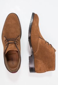 Bottes courtes en daim marron avec un design à lacets, un bout rond, et une semelle en caoutchouc noire contrastante avec une bande de roulement texturée. Marque ALDO visible à l'intérieur.