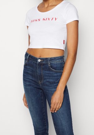 Kobiety noszące biały krótki T-shirt z napisem „Miss Sixty” oraz niebieskie, wysokiej talii jeansy, stojące na jednolitym białym tle.