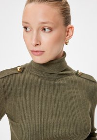 Oliwkowy sweter z golfem, prążkowany, z złotymi guzikami na ramieniu, teksturowany materiał, dopasowany krój i wysoki kołnierz.