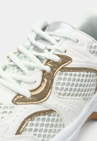 Λευκό αθλητικό παπούτσι με διχτυωτές επιφάνειες, χρυσές λεπτομέρειες και ασημένιες τρύπες για κορδόνια. Διαθέτει στρογγυλεμένη μύτη και επίπεδα κορδόνια για σταθερή εφαρμογή. Εξωτερική σόλα από υφασμένο καουτσούκ.