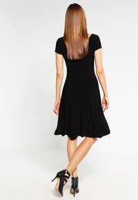 Robe noire à manches courtes avec une jupe évasée, fabriquée en tissu extensible. La vue de dos montre une silhouette fluide et un ourlet à mi-genoux. Sandales à talons hauts noires.