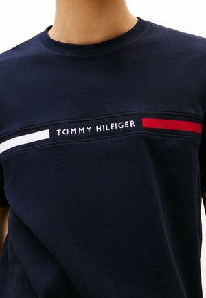 Granatowy t-shirt z logo Tommy Hilfiger umieszczonym na środku między poziomymi białymi i czerwonymi paskami na piersi.