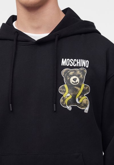 Μαύρο φούτερ με κουκούλα με λευκό κείμενο "MOSCHINO" πάνω από ένα σχέδιο με μια αρκούδα τέντι τυλιγμένη σε κίτρινη μεζούρα στο στήθος.