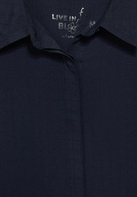 Camicia blu navy con un motivo a griglia testurizzato, colletto a punta e orlo pulito. Include un'etichetta testuale argentata con scritto "Live in Bloom".
