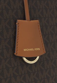 Etichetta Michael Kors in pelle marrone con lettering dorato, attaccata a un tessuto marrone scuro texturizzato con un motivo. Accento metallico circolare.
