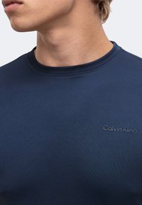 Κοντινό ενός ατόμου που φορά μπλουζάκι Calvin Klein σε σκούρο μπλε χρώμα με στρογγυλή λαιμόκοψη, δείχνοντας τον λαιμό και το κάτω μέρος του προσώπου.