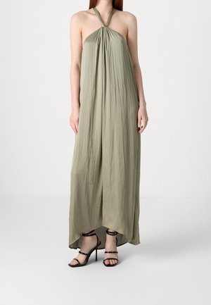 Frau trägt ein ärmelloses, olivgrünes Maxikleid mit Neckholder und Falten, kombiniert mit schwarzen, hochhackigen Riemchensandalen.