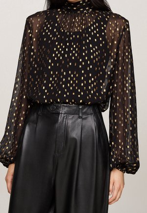 Chemisier noir transparent à manches longues avec motif de rectangles dorés, porté avec un pantalon en cuir noir taille haute sur une personne debout.