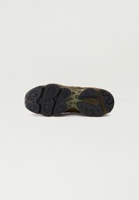 Semelle d'une chaussure New Balance marron et olive montrant des rainures en caoutchouc noir et le logo de la marque sur un fond clair uni.
