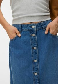 Rokas ieliktas zilos pogu priekšējā džinsu svārku kabatās, valkāta ar gaiši pelēku ribotu topu без piedurknēm.