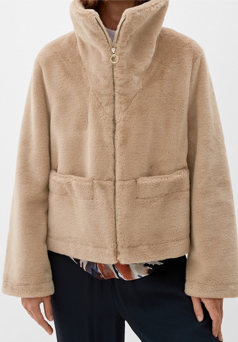 Veste beige en peluche zippée avec col montant, manches longues et détails de coutures horizontales, portée par-dessus un haut à motifs et un pantalon noir.