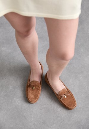 Brūni zamšādas loafers ar priekšējo banti un zelta tonī apdari, plakanā dizainā un mīkstā tekstūrā, novietoti uz neitrāla pelēka virsmas.