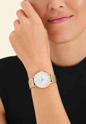 Montre en or avec un bracelet en maille métallique. Cadran rond blanc avec des index des heures en or. Portée au poignet, partiellement obscurcie par une main.