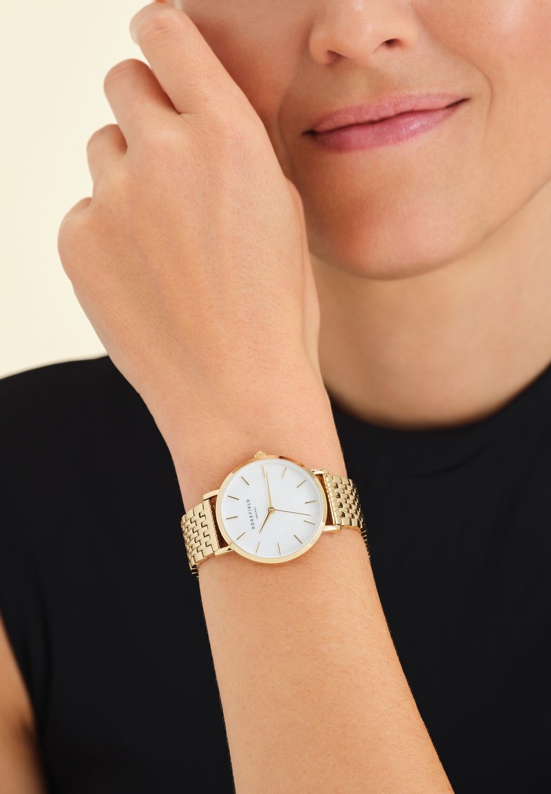Reloj de oro con una correa de malla metálica. Carátula redonda blanca con marcadores de hora dorados. Lleva puesto en una muñeca, parcialmente oscurecido por una mano.