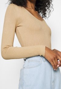 Kobieta z kręconymi włosami, ubrana w beżowy, prążkowany top z długim rękawem i jasnoniebieskie spodnie z wysokim stanem, stoi z rękami blisko talii.