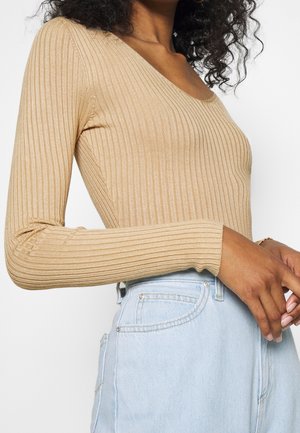 Femme aux cheveux bouclés portant un haut à manches longues en ribbed beige et un jean taille haute bleu clair, se tenant les mains près de la taille.