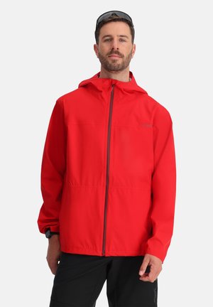 Homme portant une veste à capuche rouge vif fermée, pantalon noir et lunettes de soleil sur la tête, debout devant un fond clair uni.