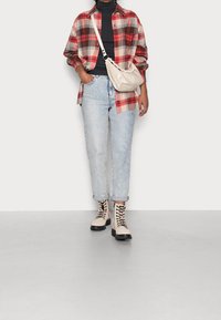 Chemise à carreaux rouge et crème portée sur un col roulé noir, jean en denim bleu clair, et bottes beiges à lacets avec semelles noires. Comprend un sac bandoulière beige.