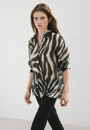 Femme portant une chemise à boutons rayée zébrée noire et blanche avec les manches retroussées et un pantalon noir, debout devant un fond uni.