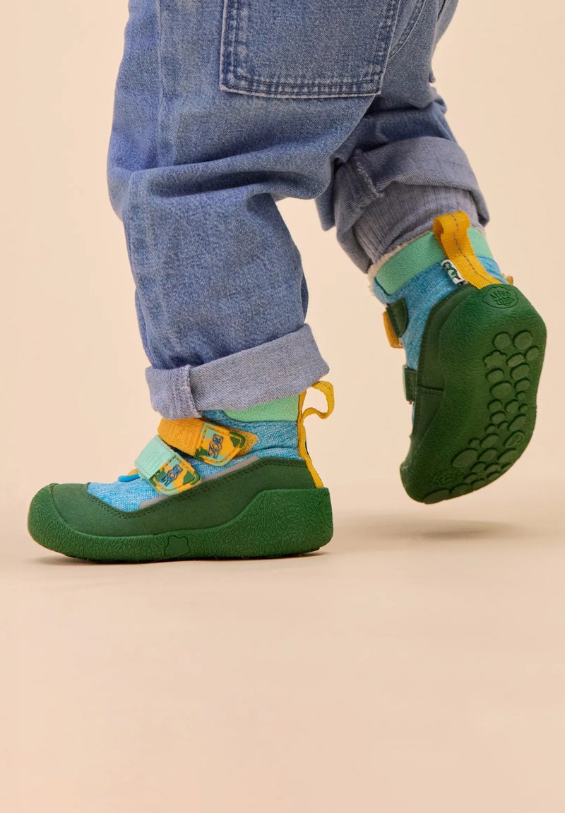 Scarpe per bambini verdi e blu con suola in gomma testurizzata, dotate di cinturini gialli, collo imbottito e dettagli a stampa giocosi.
