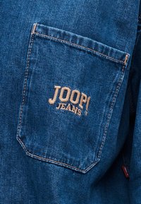 Τσέπη από τζιν ύφασμα με πορτοκαλί ραφές και μπεζ κεντητό κείμενο "JOOP! JEANS" στο κέντρο της τσέπης.