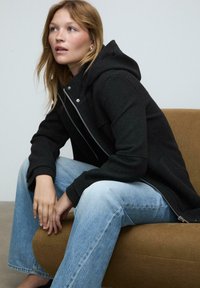 Čierna kapucňová bunda so strieborným zipsom, oblečená cez svetlomodré džínsy. Tkanina vyzerá štruktúrovaná a model sedí na hnedom gauči.