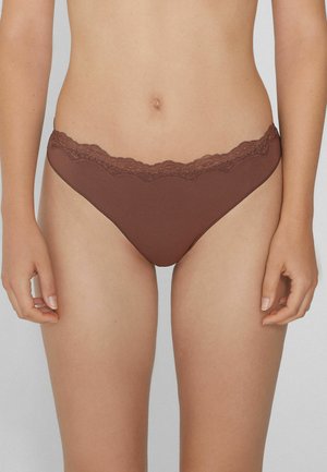 Frau in brauner Bikini-Unterwäsche mit Spitze, die vor einem schlichten hellen Hintergrund steht, mit entspannten Händen an den Seiten.
