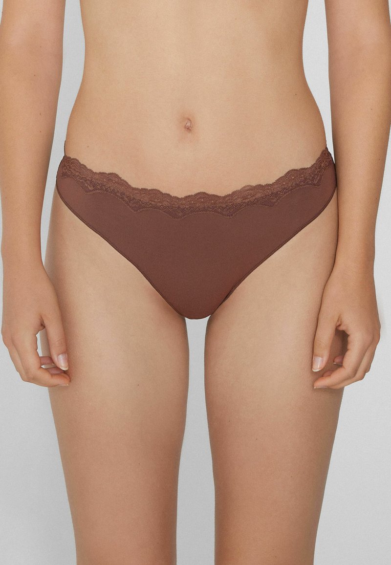 Frau in brauner Bikini-Unterwäsche mit Spitze, die vor einem schlichten hellen Hintergrund steht, mit entspannten Händen an den Seiten.