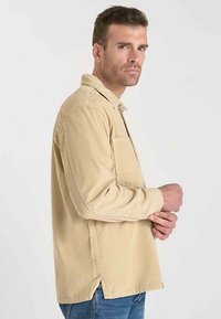 Chemise en velours côtelé beige avec col boutonné, manches longues, poche poitrine unique et fentes sur les côtés ; tissu texturé à nervures verticales.