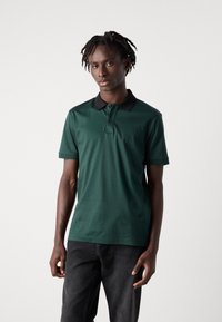 Tmavo zelená polokošeľa s čiernym golierom, krátkymi rukávmi a zapínaním na tri gombíky. Nosená s tmavými džínami, ktoré majú hladkú textúru látky.