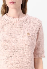 Hellrosa gestrickter Pullover mit kurzen Ärmeln, einem gerippten Rundhalsausschnitt und einer Tasche mit einem goldenen Akzentknopf. Gut sichtbare strukturiertes Gewebe.