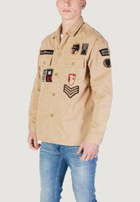 Beige bomulds skjorte med flere broderede lapper, to brystlommer og knaplukning. Lange ærmer med knapmanchetter.
