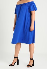 Sininen olkalta paljastava mekko, joka on valmistettu pehmeästä kankaasta, lyhyillä hihoilla ja rentolla istuvuudella. Yhdistetty mustiin paksupohjaisiin sandaaleihin.