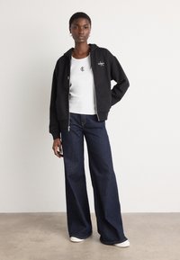 Black zip-up hoodie, white fitted t-shirt with logo, wide-leg blue denim jeans, and white sneakers. Simple design, casual look.