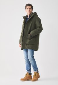 Oliven grøn polstret parka med hætte, frontlynlås, to store frontlommer, båret over en blå skjorte og lyseblå jeans. Brune arbejdsstøvler.