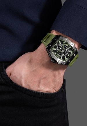 Muñeca izquierda con reloj cronógrafo negro y verde con esfera cuadrada y correa de silicona verde, mano descansando dentro del bolsillo de pantalones negros.