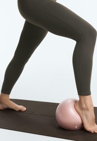 Jambes en leggings foncés, un pied sur un ballon de gym rose posé sur un tapis de yoga noir, l'autre pied à plat sur le tapis.