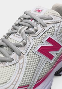 Primo piano di una sneaker New Balance bianca e beige con lacci grigi e caratteristico logo "N" rosa su tomaia in mesh e materiale sintetico.