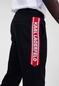 Svarta sweatpants med sidopanel som har en röd och vit "KARL LAGERFELD"-logotyp, slät textur och elastisk midja.