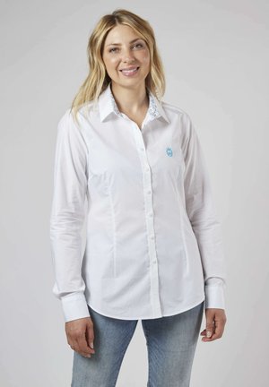 Chemise blanche à manches longues, avec un logo brodé en accent bleu et un col doublé de motifs floraux. Texture du tissu lisse.
