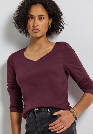 Haut bordeaux à manches longues avec un décolleté en V, fabriqué en tissu doux, associé à un jean noir et un bracelet texturé en or.