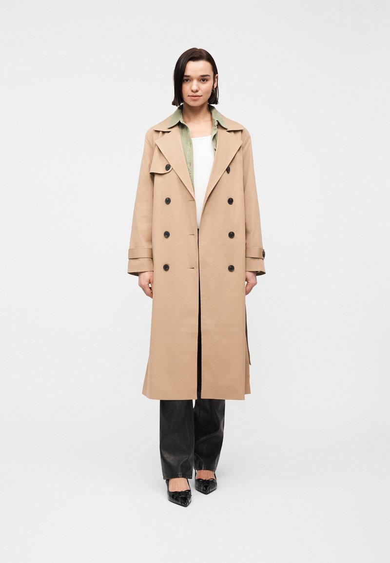 Femme debout, portant un trench-coat beige à double boutonnage, une chemise verte, un haut blanc, un pantalon noir et des chaussures noires pointues sur un fond blanc.