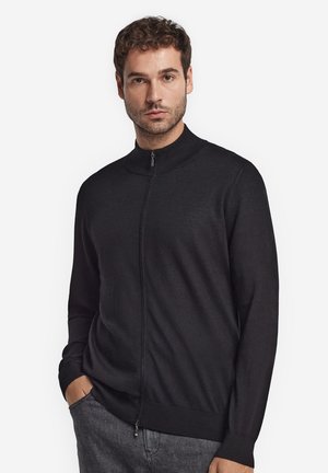 Homme portant un pull zippé noir à col montant et un pantalon gris, debout avec une main dans la poche sur un fond uni.
