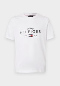 Bílé tričko s krátkým rukávem s textem "Tommy Hilfiger 1985" a červenou, bílou a černou vlajkou loga Tommy Hilfiger na přední straně.
