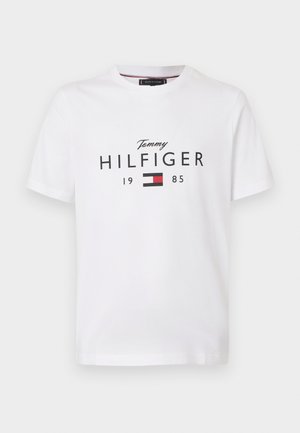 Bílé tričko s krátkým rukávem s textem "Tommy Hilfiger 1985" a červenou, bílou a černou vlajkou loga Tommy Hilfiger na přední straně.