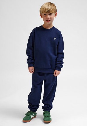 Marineblauwe sweatshirt en bijpassende joggers van zachte stof. Het shirt heeft een klein logo op de borst; de broek heeft elastische manchetten. Groene schoenen.