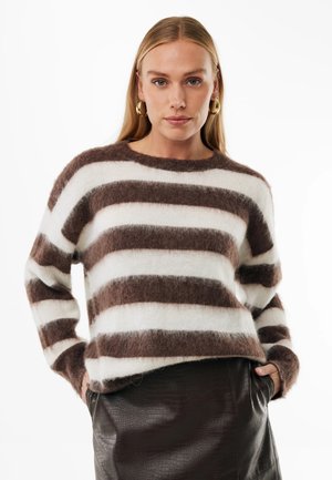 Kvinde med langt blond hår iført guldringeøreringe, en fluffy brun og hvid stribet sweater og et mørkt tekstureret skørt med hænderne i lommerne.