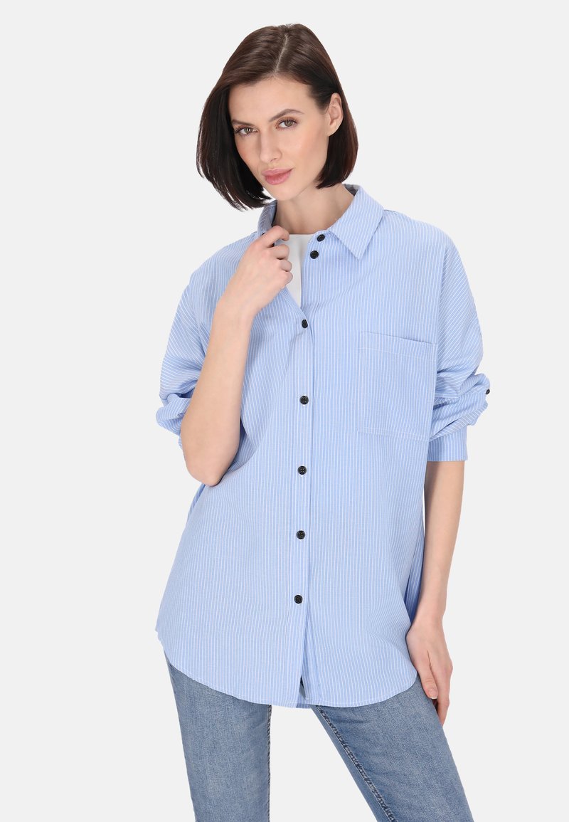 Camicia button-up a strisce blu con colletto, taschino sul petto, e bottoni neri, abbinata a jeans blu aderenti. Tessuto morbido.