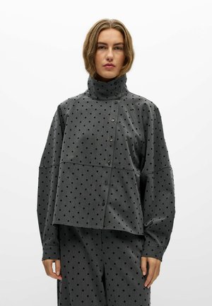 Un mannequin se tient devant un fond blanc, vêtu d’un ensemble à pois gris et noirs, avec un col montant et une veste boutonnée sur le devant.